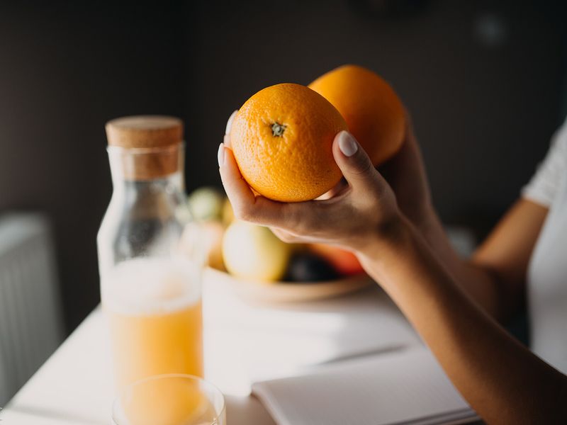 A hand lifting an orange from the fruit bowl on a sunlit breakfast table, beside a jug of juice and an open notebook